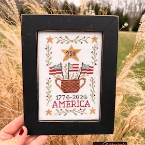 May include: Framed cross-stitch art with a black frame. The design features American flags, stars, and the text "1776-2026 AMERICA" and "250". The artwork is in a rustic style, celebrating a patriotic theme.