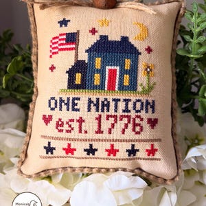 May include: A cross-stitched patriotic ornament featuring a blue house with a red door and an American flag. The text "ONE NATION est. 1776" is stitched below the house. The ornament is trimmed with a jute rope and topped with a small bell.