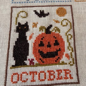 May include: A cross-stitch embroidery on light beige fabric featuring a black cat and an orange jack-o'-lantern. Above the pumpkin is a black bat and a yellow moon. The word OCTOBER is stitched in orange at the bottom, all within a brown border.