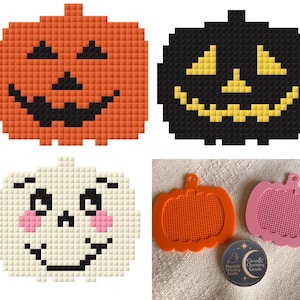 May include: Three pixel-art style pumpkin designs: one orange with black features, one black with yellow features, and one white with black features and pink cheeks. Also, two plastic pumpkin-shaped cross-stitch blanks in orange and pink.