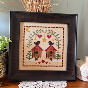 May include: Framed cross-stitch artwork featuring two birdhouses with birds, hearts, and stars. The artwork is in a dark wooden frame and rests on a wooden surface. The cross-stitch design includes brown, red, green, and white details, with a beige background.