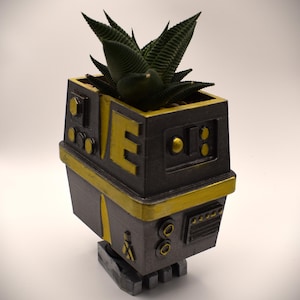May include: A dark gray and yellow planter shaped like a robotic head with mechanical details, including circular buttons and rectangular panels. A dark green succulent plant with pointed, textured leaves is planted inside the top opening.