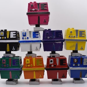 以下が含まれることがあります： カラフルな手作りのロボットフィギュアのコレクション。各ロボットは長方形のボディと円筒形のベースを持ち、ピンク、黒、白、青、紫、黄色、オレンジ、赤、緑など様々な色があります。ロボットは積み重ねられた形で配置されています。