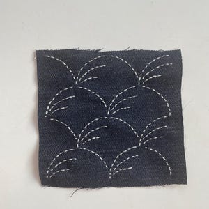 May include: A square of dark blue denim fabric with a repeating pattern of white stitched arcs. The arcs are arranged in a grid-like fashion, creating a textured surface. The edges of the fabric are raw and unfinished.