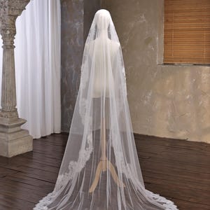 May include: A long, sheer white bridal veil with a delicate lace trim. The veil is displayed on a mannequin, showcasing its flowing design. The lace trim adds a touch of elegance to the wedding accessory.