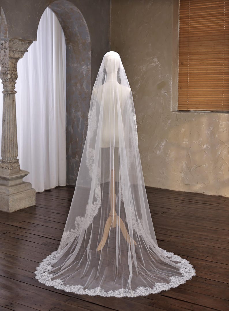 May include: A long, sheer white bridal veil with a delicate lace trim. The veil is displayed on a mannequin, showcasing its flowing design. The lace trim adds a touch of elegance to the wedding accessory.