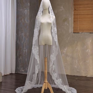 May include: A long, white bridal veil with intricate lace detailing along the edges. The veil is displayed on a mannequin, showcasing its flowing design. The lace pattern adds a delicate touch to the sheer fabric, creating an elegant look.