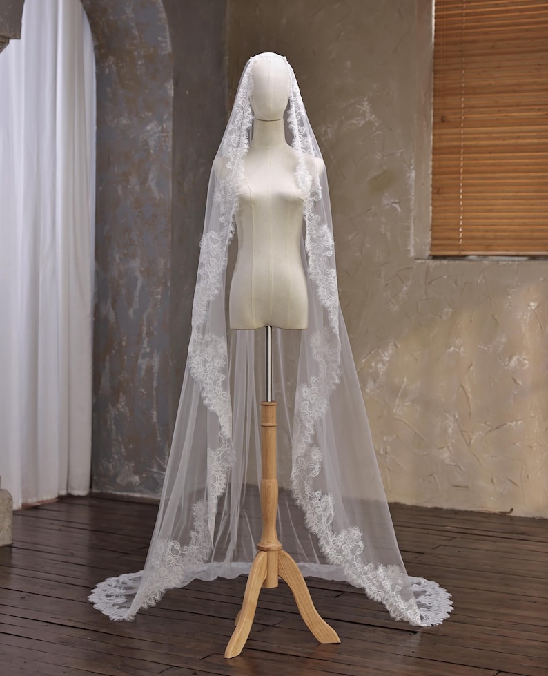 May include: A long, white bridal veil with intricate lace detailing along the edges. The veil is displayed on a mannequin, showcasing its flowing design. The lace pattern adds a delicate touch to the sheer fabric, creating an elegant look.