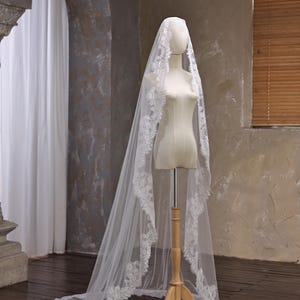 May include: A white bridal veil with a delicate lace trim is draped over a mannequin. The veil is long, flowing, and has a train. The mannequin is on a wooden stand, set against a neutral background.