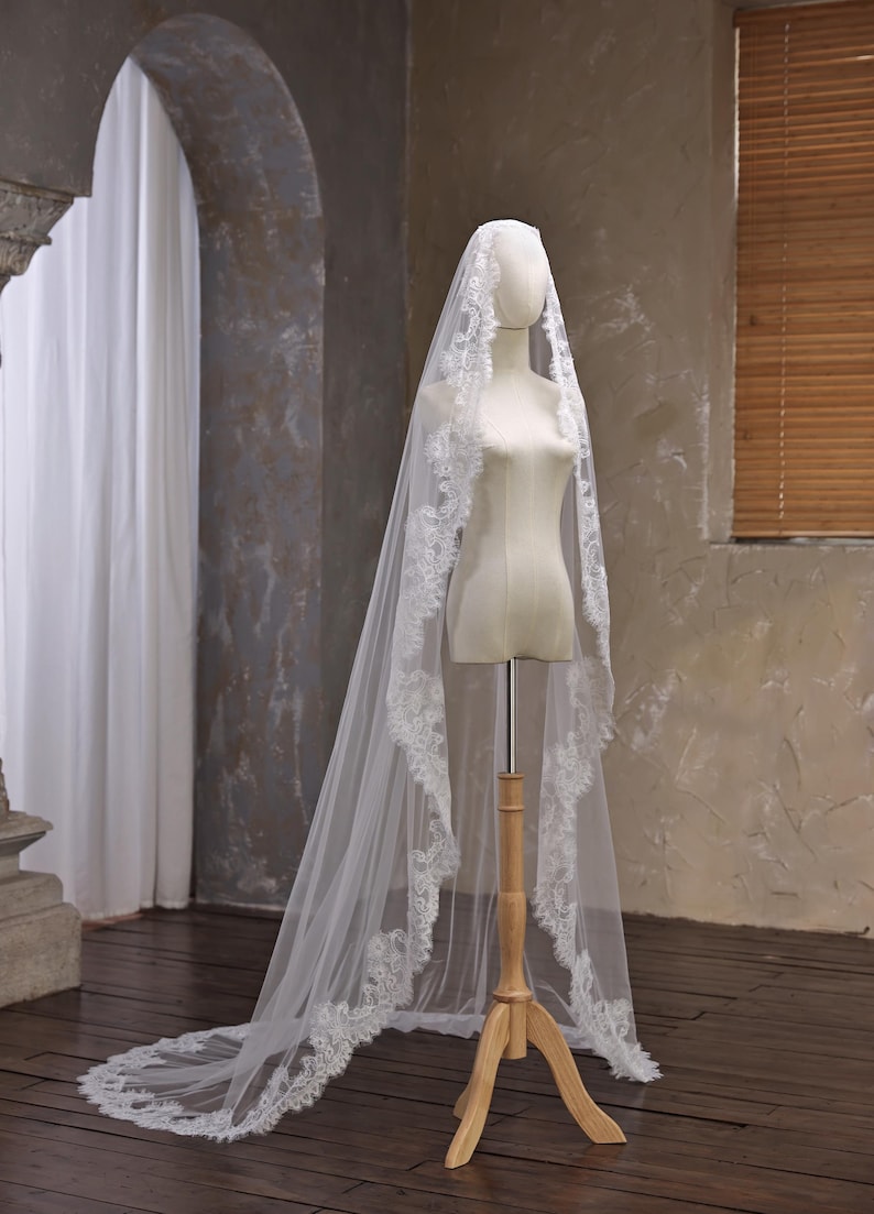 May include: A white bridal veil with a delicate lace trim is draped over a mannequin. The veil is long, flowing, and has a train. The mannequin is on a wooden stand, set against a neutral background.