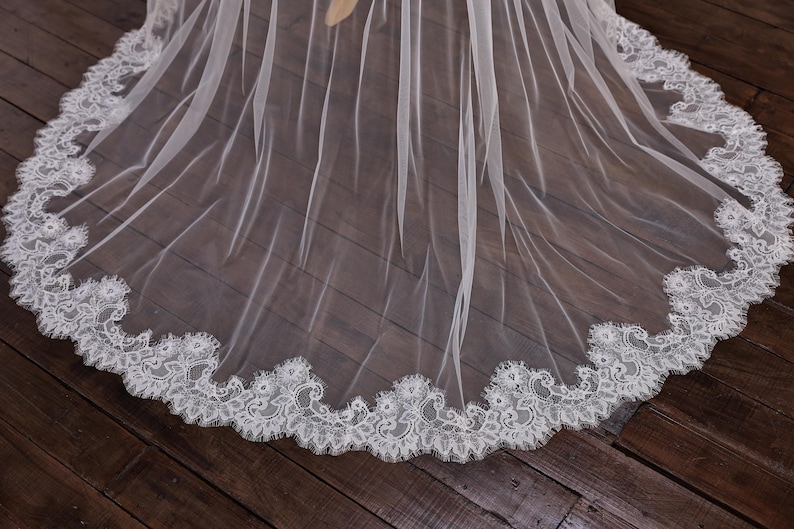 May include: A sheer white wedding veil with a delicate, scalloped lace trim. The veil is made of a lightweight fabric that drapes gracefully. The lace trim features an intricate floral design, adding a touch of elegance to the bridal accessory.