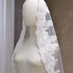 Puede incluir: Un velo de novia blanco con un delicado ribete de encaje floral. El velo está drapeado sobre un maniquí, mostrando su tejido transparente y su intrincado diseño. El ribete de encaje añade un toque de elegancia.