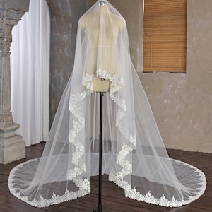 May include: A long, flowing white bridal veil with a detailed lace trim, displayed on a mannequin. The veil has intricate floral lace detailing along the edges. The mannequin is on a black stand.