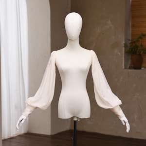 May include: A cream-colored long-sleeve top displayed on a mannequin. The garment features a fitted bodice and sheer, voluminous bishop sleeves that gather at the wrists with fitted cuffs. The fabric is smooth and opaque on the torso.