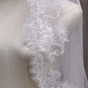 May include: A white bridal veil with a delicate lace trim. The lace features a floral pattern with scalloped edges. The veil is draped over a mannequin, showcasing the intricate details and sheer fabric.