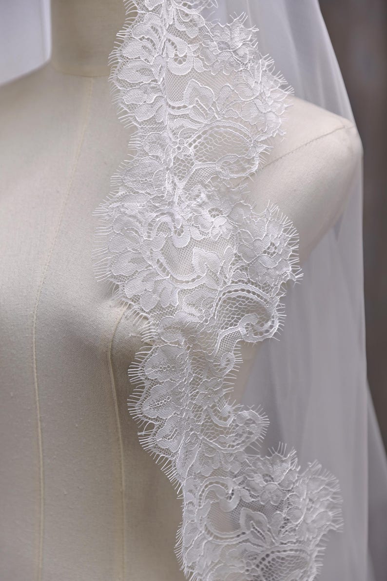 May include: A white bridal veil with a delicate lace trim. The lace features a floral pattern with scalloped edges. The veil is draped over a mannequin, showcasing the intricate details and sheer fabric.