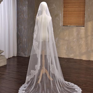 May include: A long, white bridal veil with delicate lace trim. The veil is draped over a mannequin, showcasing its length and intricate design. The lace detailing is visible along the edges, adding a touch of elegance to the wedding accessory.