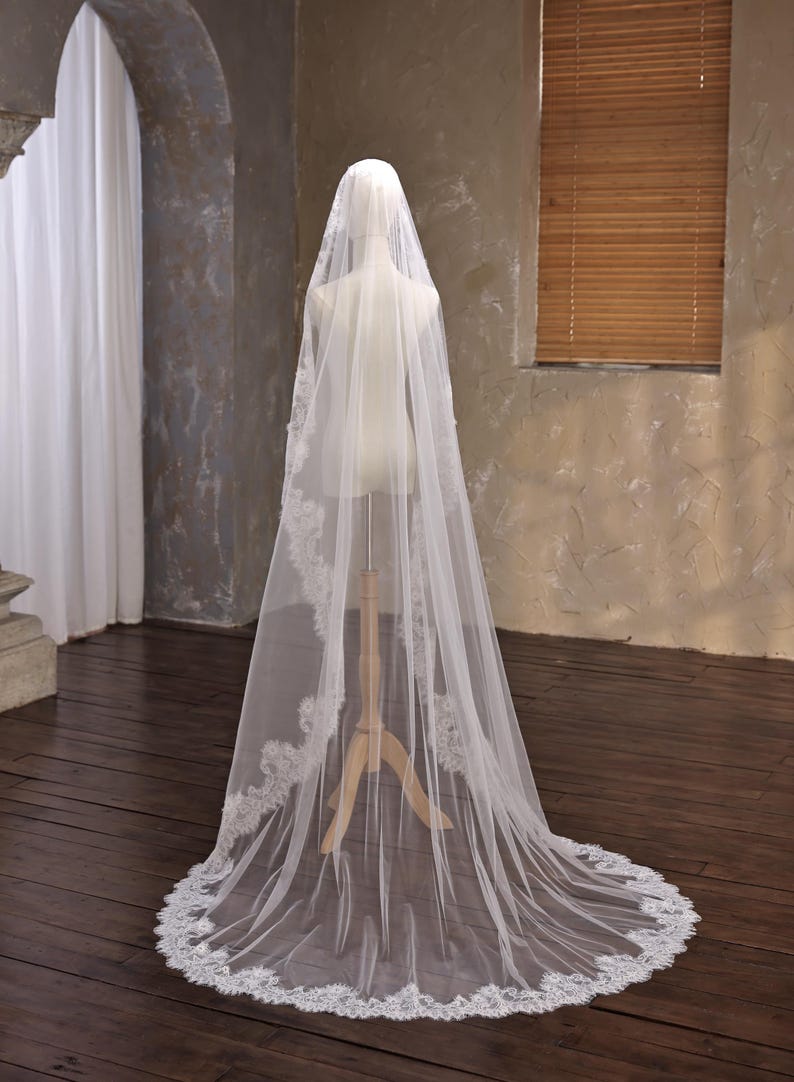 May include: A long, white bridal veil with delicate lace trim. The veil is draped over a mannequin, showcasing its length and intricate design. The lace detailing is visible along the edges, adding a touch of elegance to the wedding accessory.