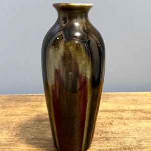 May include: A small, glossy vase with a narrow neck and a flared rim. The vase has a dark brown and olive green glaze with a crackle effect. The vase is displayed on a wooden surface.