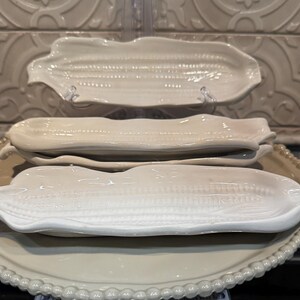 May include: A stack of ceramic serving dishes in the shape of corn on the cob. The dishes are various shades of white and cream, with textured details mimicking corn kernels. A large, round, cream-colored serving platter with a beaded edge is at the bottom.