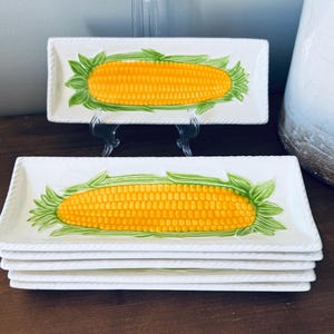 May include: A stack of rectangular white ceramic serving platters with a yellow corn design. Each platter has a rope-like border and a painted corn image with green leaves. The corn measures approximately 8 inches long.