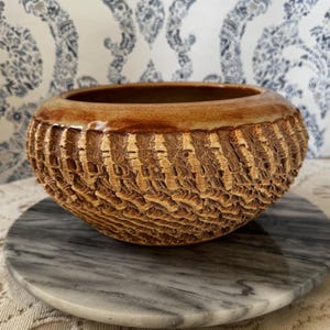 May include: A handcrafted, textured ceramic bowl with a warm, earthy color palette. The bowl features a detailed, carved pattern on its exterior, creating a unique, tactile surface. The rim is a darker shade of brown, and the interior is a lighter brown.
