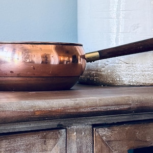 May include: A copper-colored saucepan with a dark wooden handle and a brass fitting. The small pan is round with a curved body. The handle is attached with a brass fitting. The pan is sitting on a wooden surface.