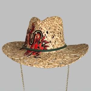 May include: A straw cowboy hat with a decorative dragon design. The hat features a woven straw construction, a green leather band, and a gold chain. The dragon is embroidered in red, black, and gold thread.