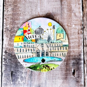 May include: A circular coaster with a watercolour illustration of Vienna, Austria. The design includes landmarks, the city name, and a Ferris wheel. The coaster is set against a wooden background.