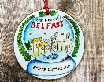 May include: A round, blue and white Christmas ornament with a snow globe design. The globe depicts a stylized illustration of the city of Belfast, Northern Ireland. The ornament has a red ribbon and the text "Our wee city Belfast" and "Merry Christmas".