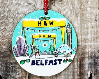 May include: A round, wooden Christmas ornament with a blue and white winter scene. The ornament features a snowman, a fish, a building, and a clock tower. The text "H&W BELFAST" is printed on the ornament.