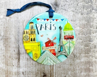 May include: A round, blue and white ceramic ornament with a colorful illustration of Parisian landmarks, including the Eiffel Tower, the Arc de Triomphe, and the Moulin Rouge. The ornament has a blue ribbon attached and the word "PARIS" in red letters.