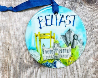 May include: A round, blue and white ornament with a hand-painted illustration of the city of Belfast, Northern Ireland. The ornament features a yellow crane, a building with a green dome, and a blue and gray building. The text "BELFAST" is written in black across the top of the ornament.
