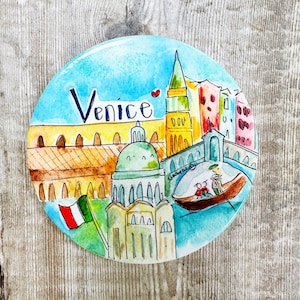 May include: A round ceramic plate with a watercolor painting of Venice, Italy. The plate features a blue sky, a yellow building with arches, a green domed building, a tall red and yellow tower, and a gondola with two people in it. The words "Venice" are written in black on the plate.