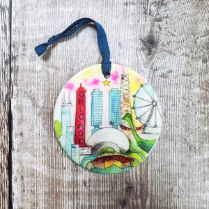 May include: A round, white ceramic ornament with a watercolor painting of the Chicago skyline. The ornament features iconic landmarks like the Willis Tower, the John Hancock Center, and the Cloud Gate sculpture. The text "CHICAGO" is written vertically on the side of the ornament.