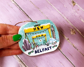 May include: A ceramic ornament featuring a blue and green illustration of a building with the text "H&W" and "BELFAST". The ornament is decorated with a snowman, a fish, and a clock tower.