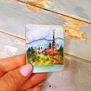 Può includere: Un dipinto quadrato ad acquerello del lago di Bled, in Slovenia, raffigurante una chiesa con un alto campanile su un'isola. L'opera d'arte include il testo "Lake Bled" ed è ambientata su uno sfondo di montagne blu e alberi verdi.