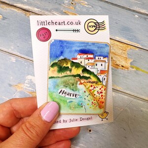 May include: A watercolor painting of Algarve, Portugal, on a white card. The artwork depicts a coastal scene with white buildings, green hills, and blue water. The card has the text "littleheart.co.uk" and "Algarve".