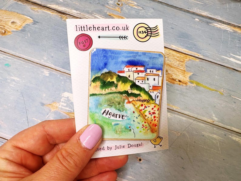 May include: A watercolor painting of Algarve, Portugal, on a white card. The artwork depicts a coastal scene with white buildings, green hills, and blue water. The card has the text "littleheart.co.uk" and "Algarve".