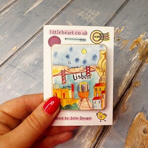 May include: A colorful illustrated magnet featuring a cityscape of Lisbon, Portugal. The magnet depicts a red bridge, a yellow tram, and buildings with red roofs. The text "Lisbon" is written in white on a red background.