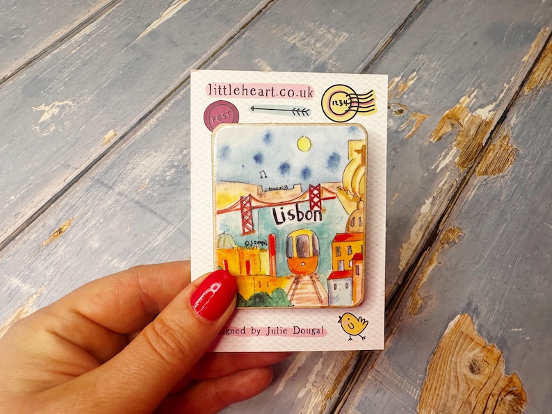 May include: A colorful illustrated magnet featuring a cityscape of Lisbon, Portugal. The magnet depicts a red bridge, a yellow tram, and buildings with red roofs. The text "Lisbon" is written in white on a red background.