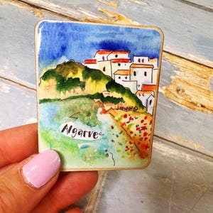 May include: A square watercolor painting of the Algarve coast. The artwork depicts white buildings with red roofs on a hillside, a blue sky, and the word "Algarve". The painting is held in a hand.