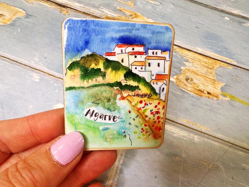 May include: A square watercolor painting of the Algarve coast. The artwork depicts white buildings with red roofs on a hillside, a blue sky, and the word "Algarve". The painting is held in a hand.