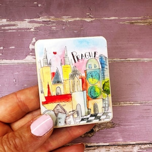 Può includere: Un'illustrazione colorata di Praga, Repubblica Ceca, con un tetto rosso, una torre dell'orologio e edifici alti. L'illustrazione è su un magnete di legno.