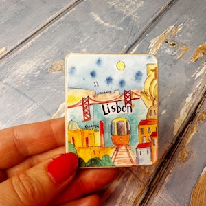 May include: A colorful magnet featuring a cartoon illustration of Lisbon, Portugal. The magnet depicts a red bridge, a yellow tram, and buildings with red roofs. The text "Lisbon" is written in a blue font.
