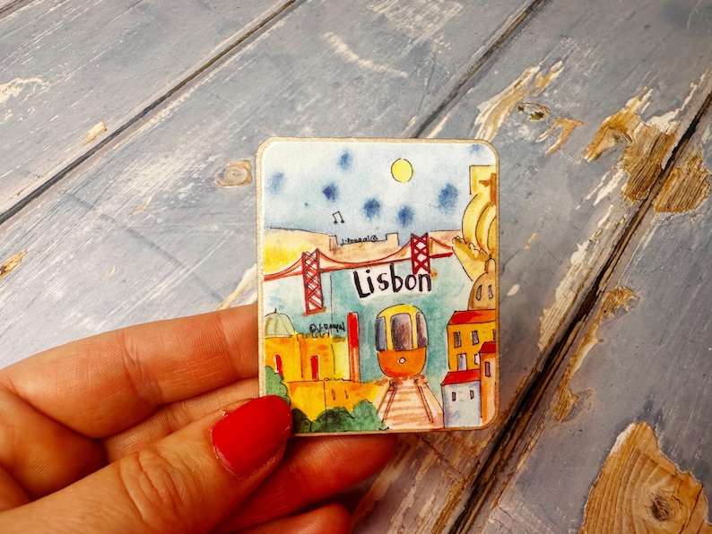 May include: A colorful magnet featuring a cartoon illustration of Lisbon, Portugal. The magnet depicts a red bridge, a yellow tram, and buildings with red roofs. The text "Lisbon" is written in a blue font.