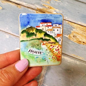 May include: A rectangular watercolor painting of Algarve, Portugal. The artwork depicts white buildings with red roofs on a green hillside, a blue sky, and the word "Algarve" in cursive.