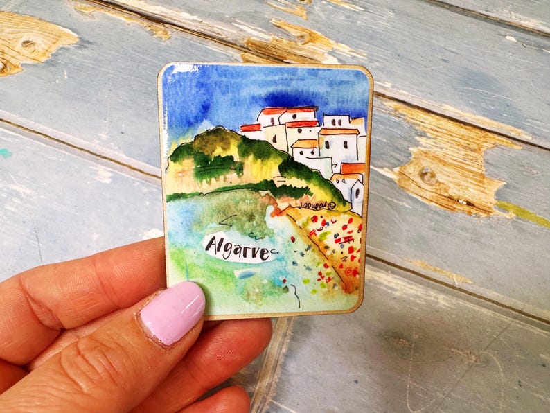 May include: A rectangular watercolor painting of Algarve, Portugal. The artwork depicts white buildings with red roofs on a green hillside, a blue sky, and the word "Algarve" in cursive.