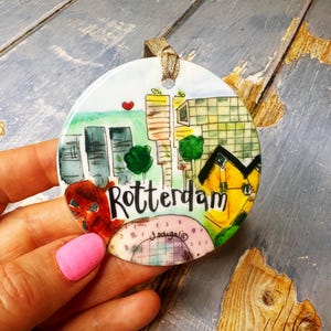 Könnte beinhalten: Runder Keramikschmuck mit einem Aquarellgemälde der Skyline von Rotterdam. Das Design umfasst Gebäude, Bäume und den Namen der Stadt in schwarzer Schrift. Ein Band ist zum Aufhängen befestigt.