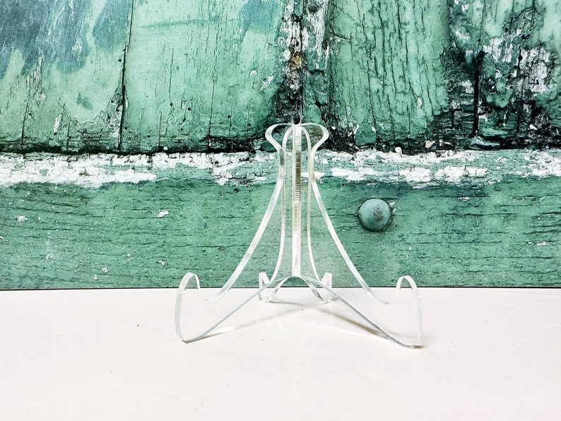 May include: A clear acrylic display stand with three legs. The stand is designed to hold a plate or other flat object.
